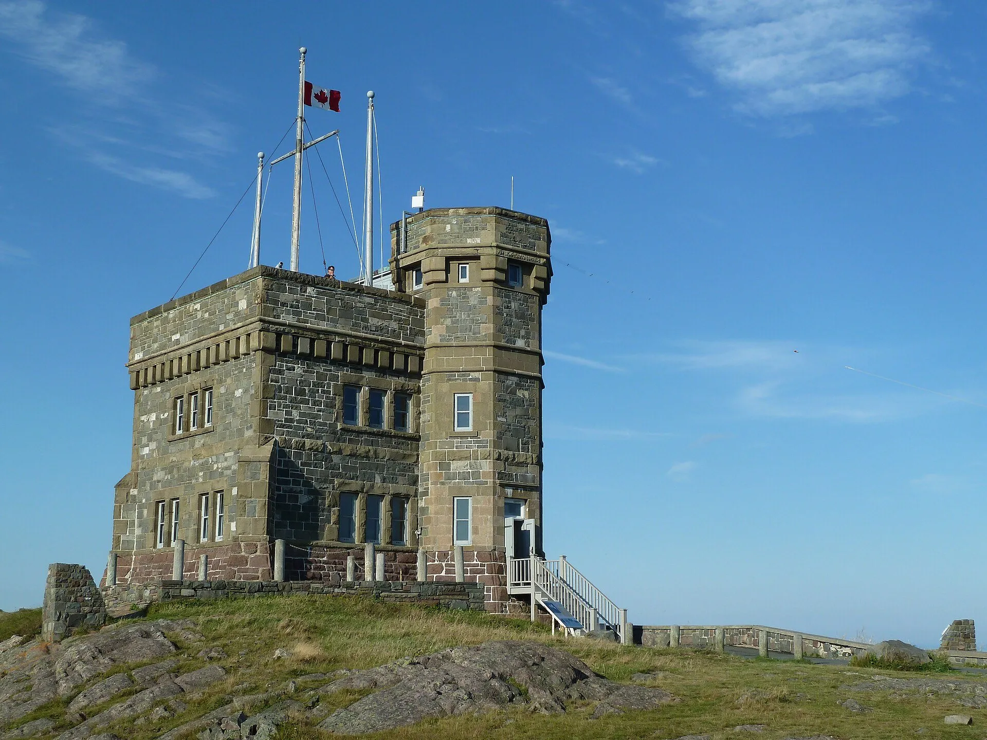 Cabot Tower, a stone castle-like tower with a Canadian flag, on the rocky summit of Signal Hill under a blue sky.