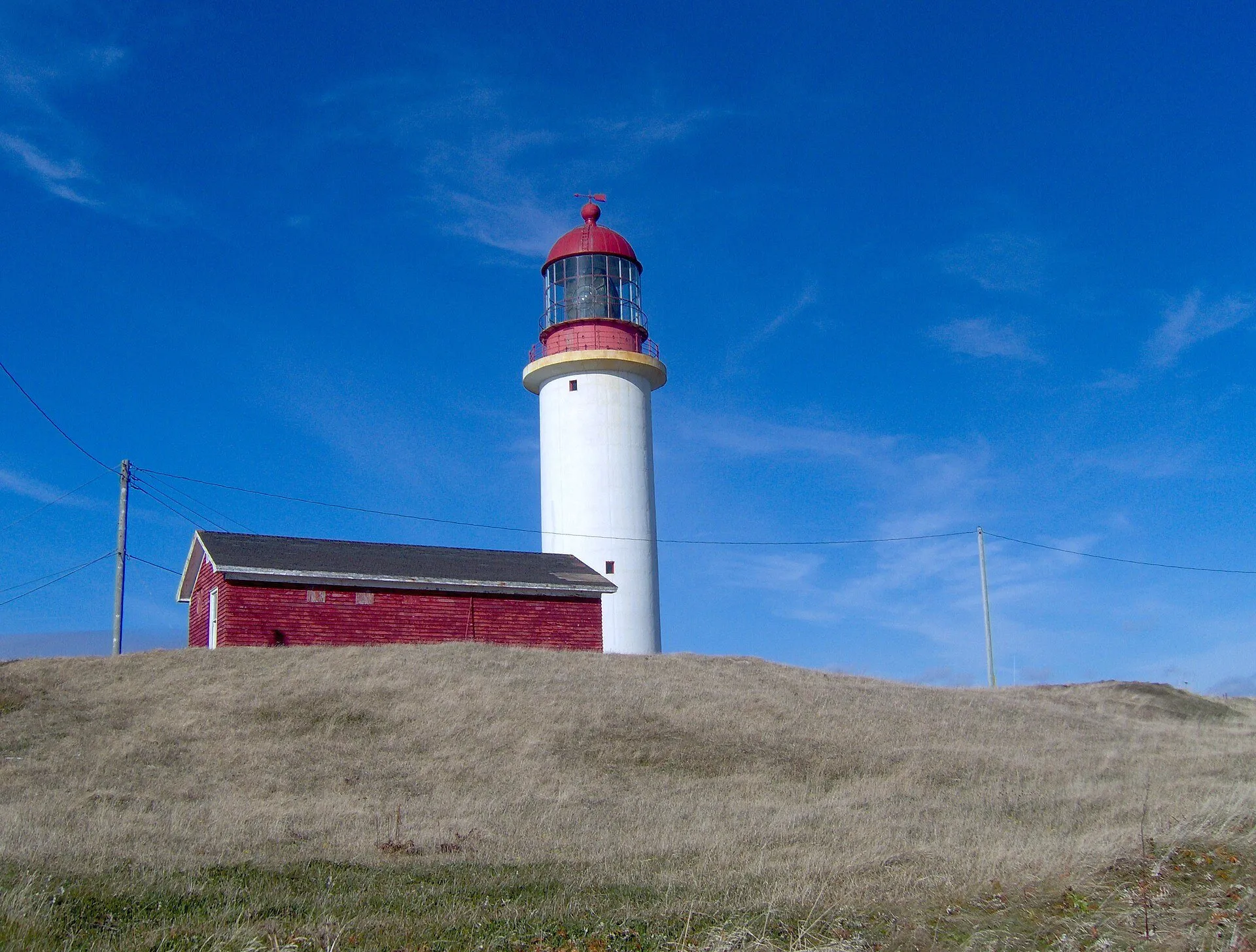 The white Cape Race lighthouse tower with its red lantern rises behind a red outbuilding on a grassy rise.