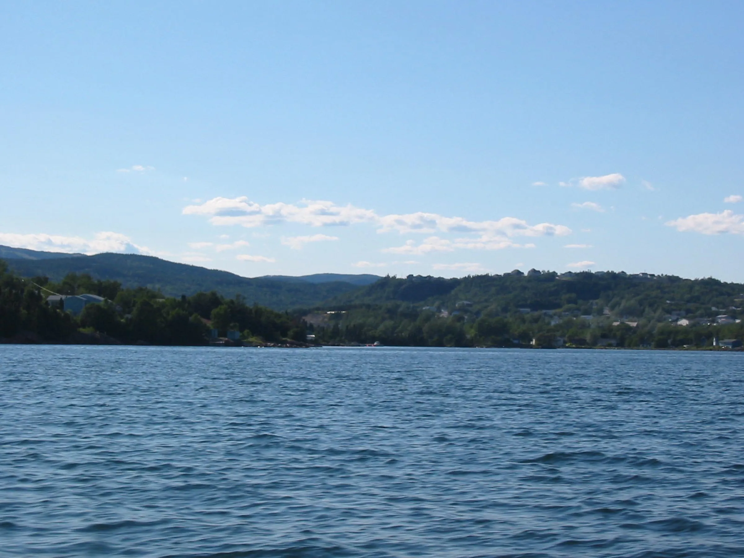 Clarenville seen from the water, the town spread along the wooded hillside above Random Sound under a clear sky.