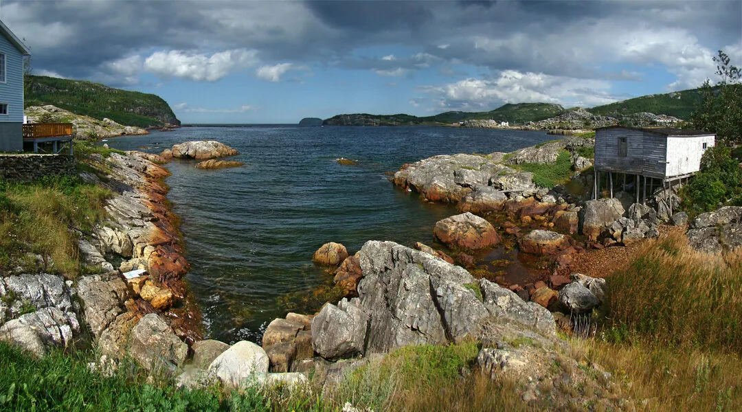 A rocky inlet at Salvage with a white fishing stage on the shore and wooded hills beyond, under a cloudy sky.