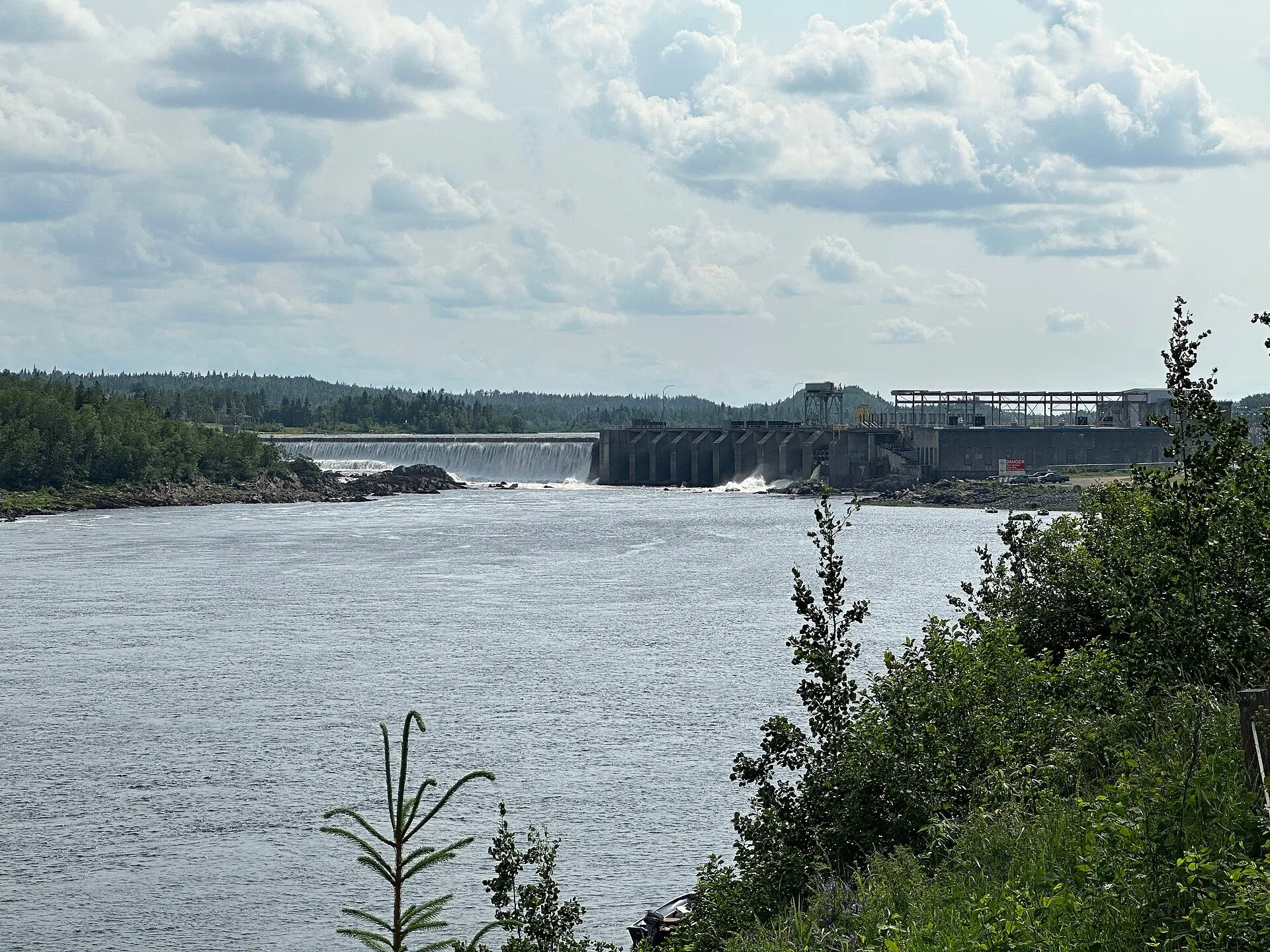 The Exploits River runs wide below the dam and power station at Bishop's Falls, framed by trees on the bank.