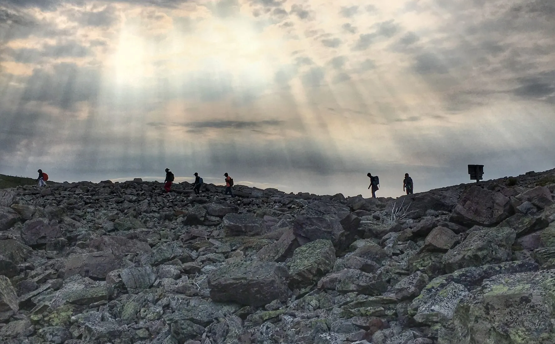 Hikers in silhouette crossing the bare boulder field on the summit of Gros Morne Mountain, sun breaking through cloud.