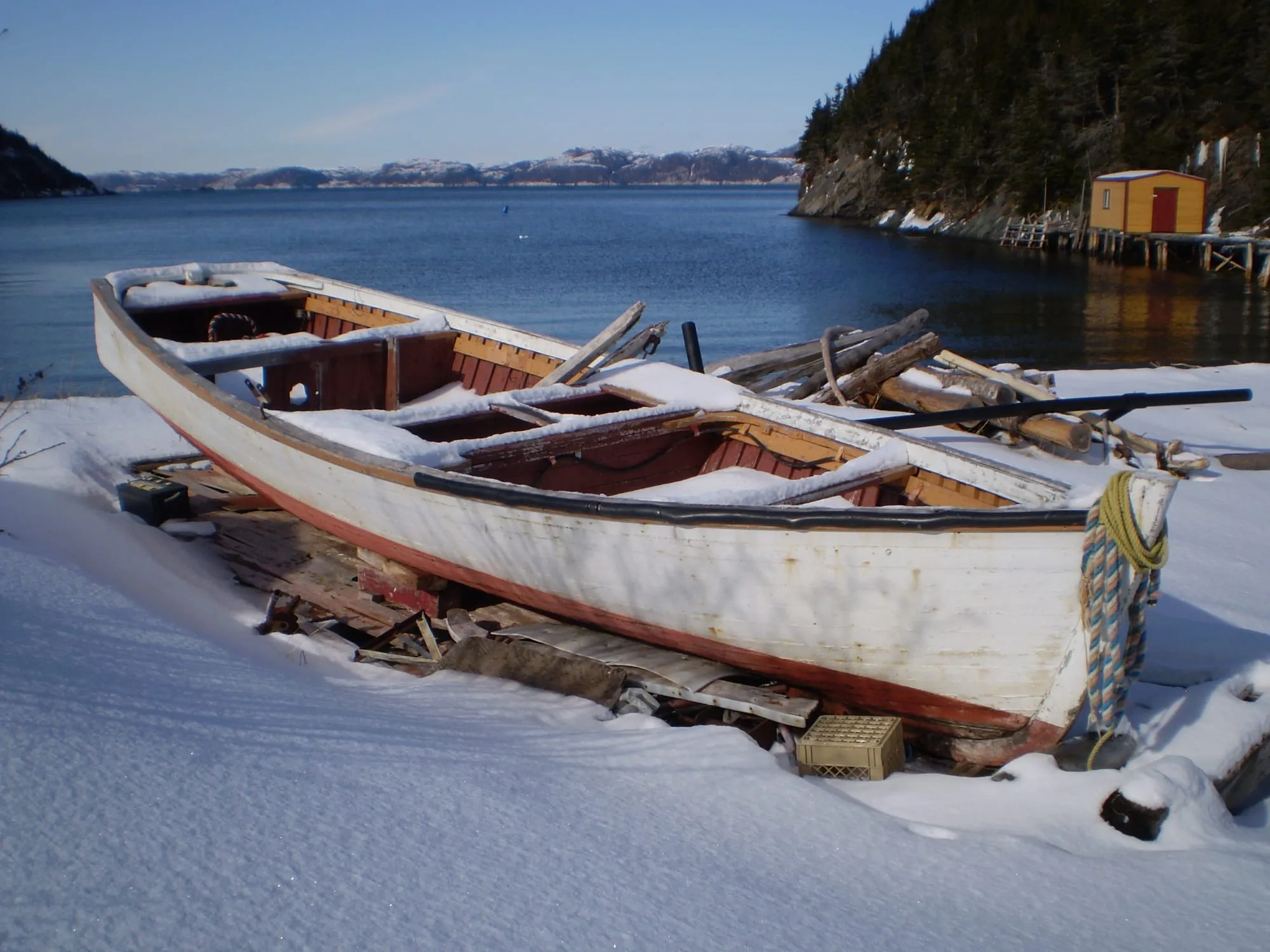 A wooden fishing boat hauled up in the snow at Hermitage on the south coast, the harbour and a wooded headland behind.