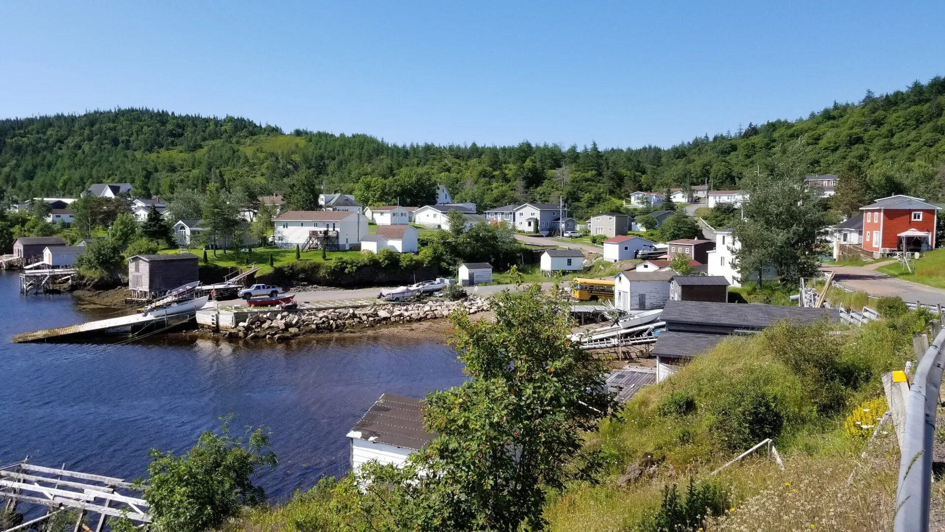 Houses, sheds and slipways along the inner harbour of Pool's Cove, with forested hills behind.