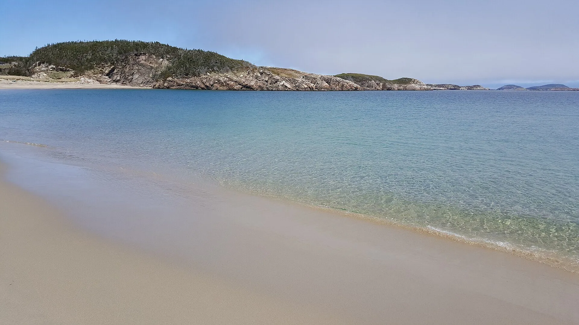 Clear shallow water over pale sand at Sandbanks Provincial Park, with a wooded headland across the bay.