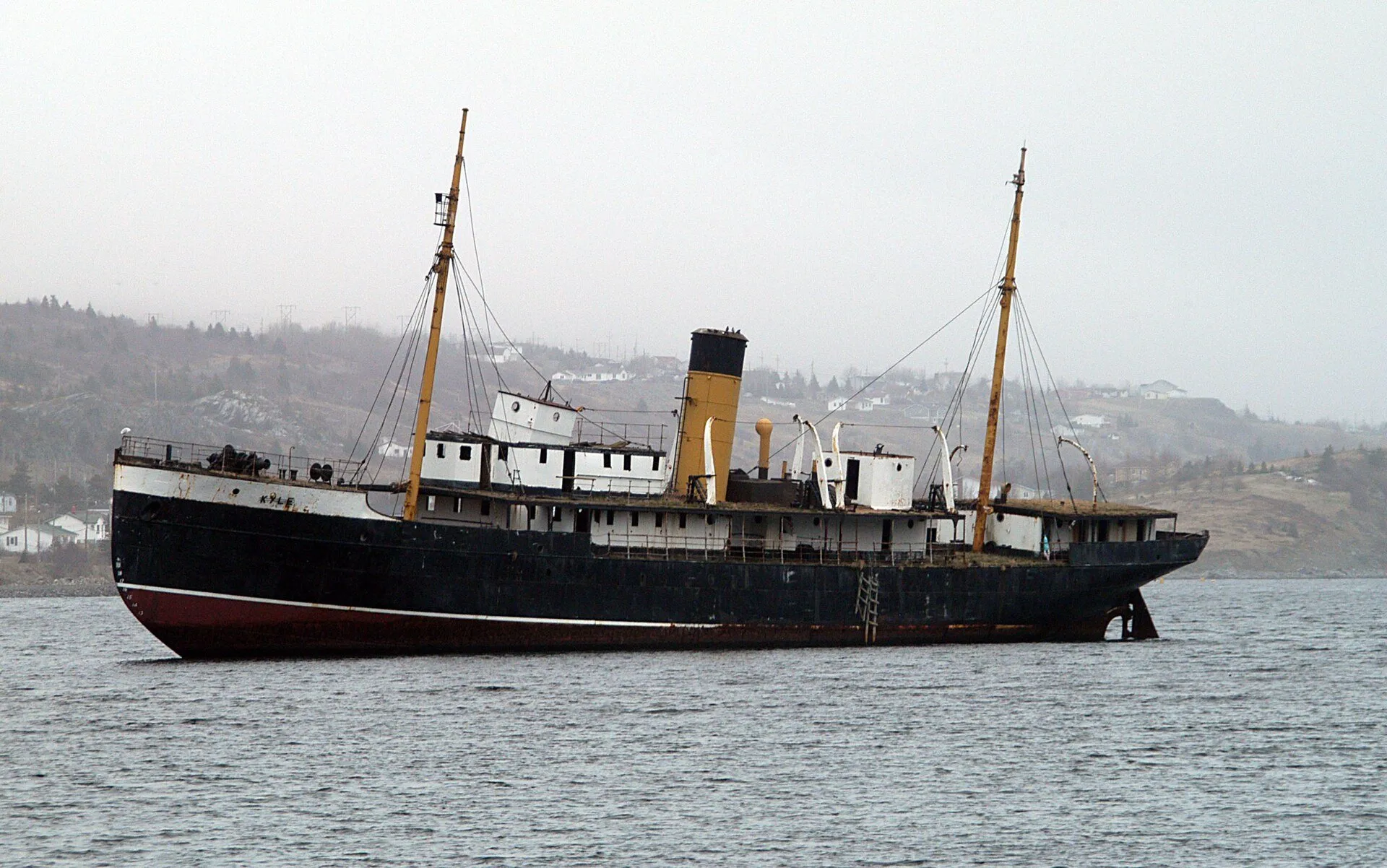 The black-hulled steamship SS Kyle sitting aground in Harbour Grace harbour, with its two masts and yellow funnel against the hills.