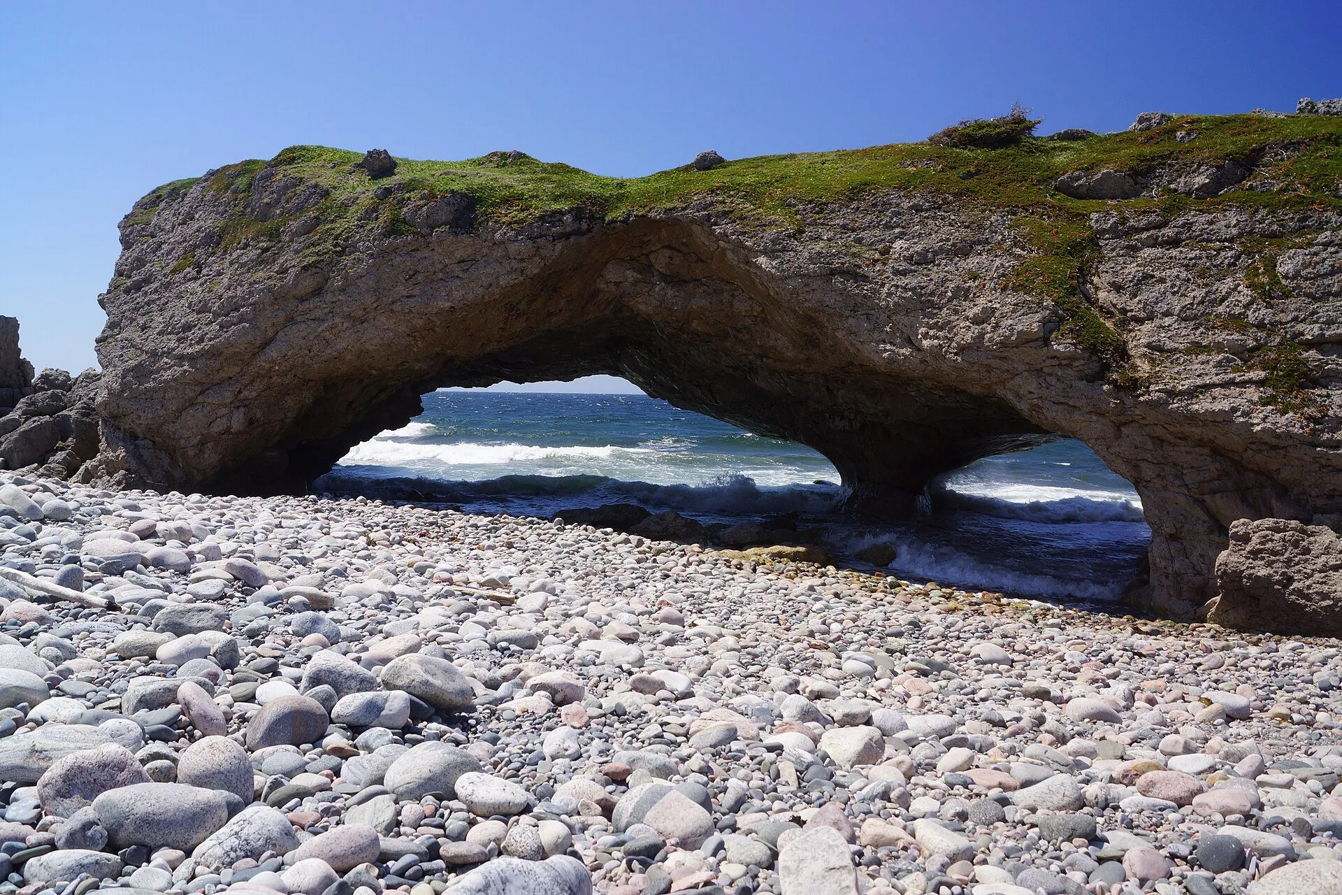The rock arches at The Arches Provincial Park, a grass-topped limestone formation with sea showing through its openings above a cobble beach.