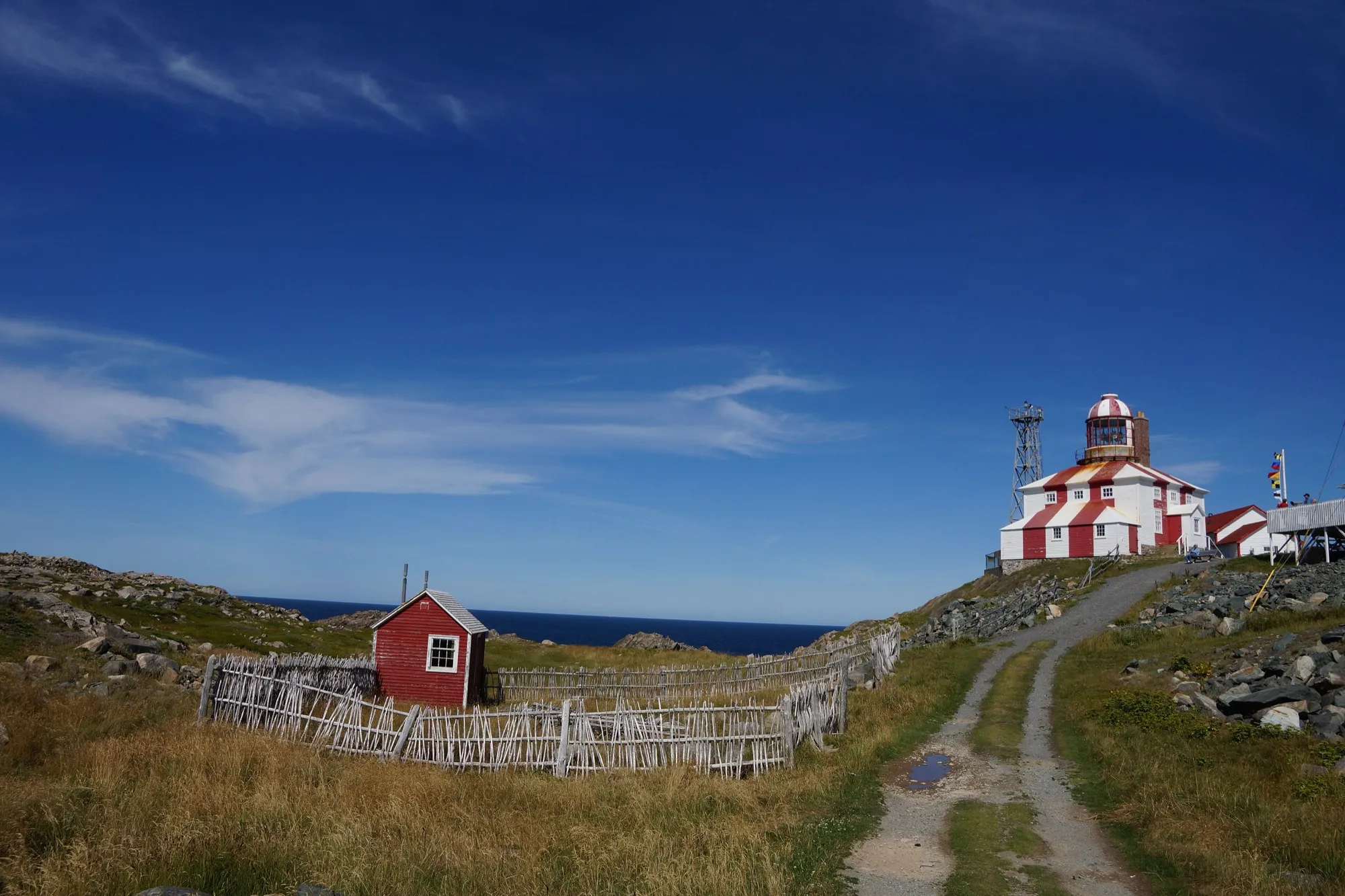 Wider view of the lighthouse up the gravel path with ocean and blue sky.
