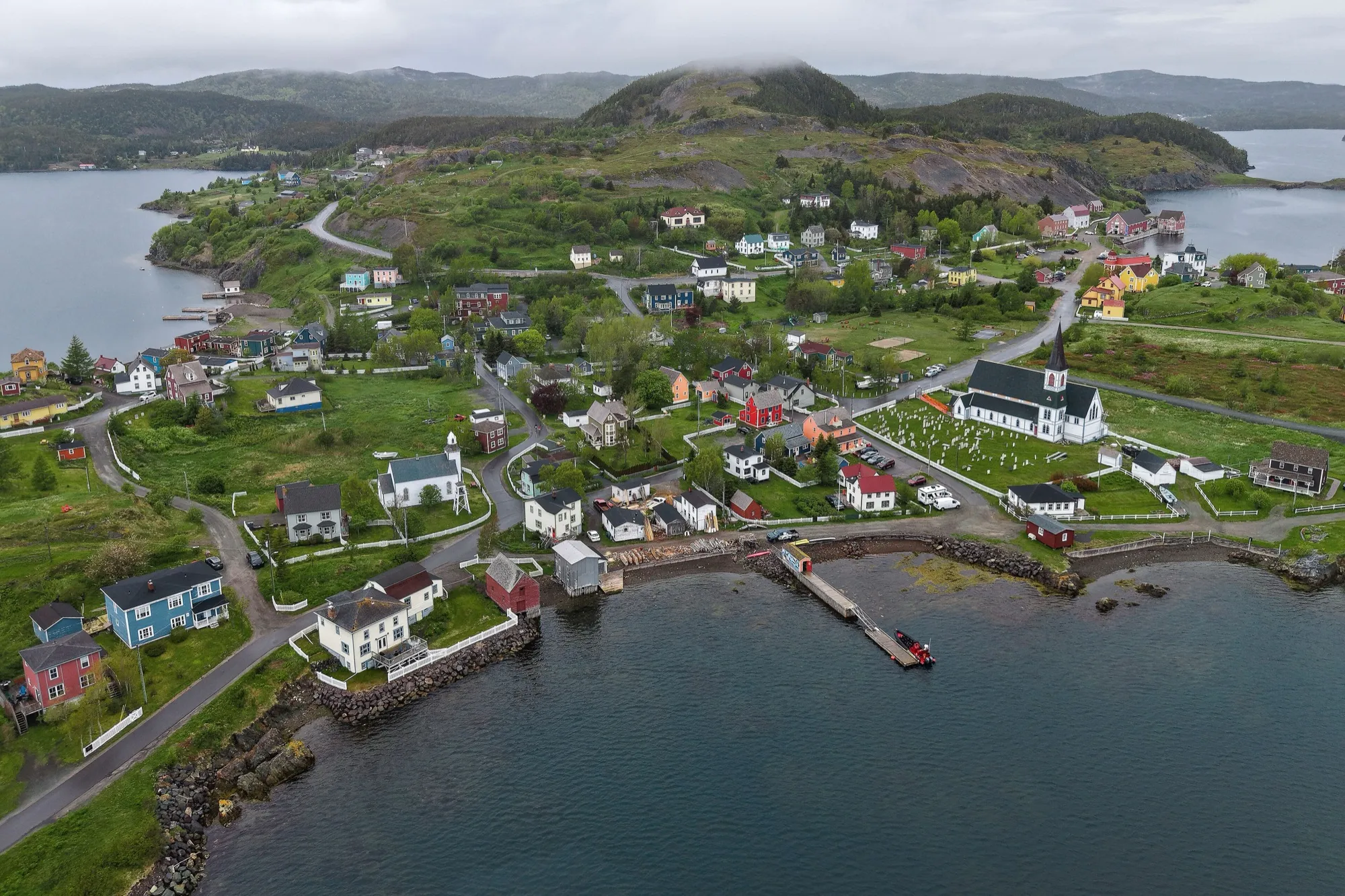 Aerial view of Trinity: coloured houses, the Anglican church, harbour and wharf, overcast summer.